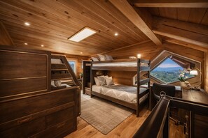 Ein Holzblockhaus-Innenraum mit Etagenbett, Schreibtisch und Blick durch ein Dachfenster.