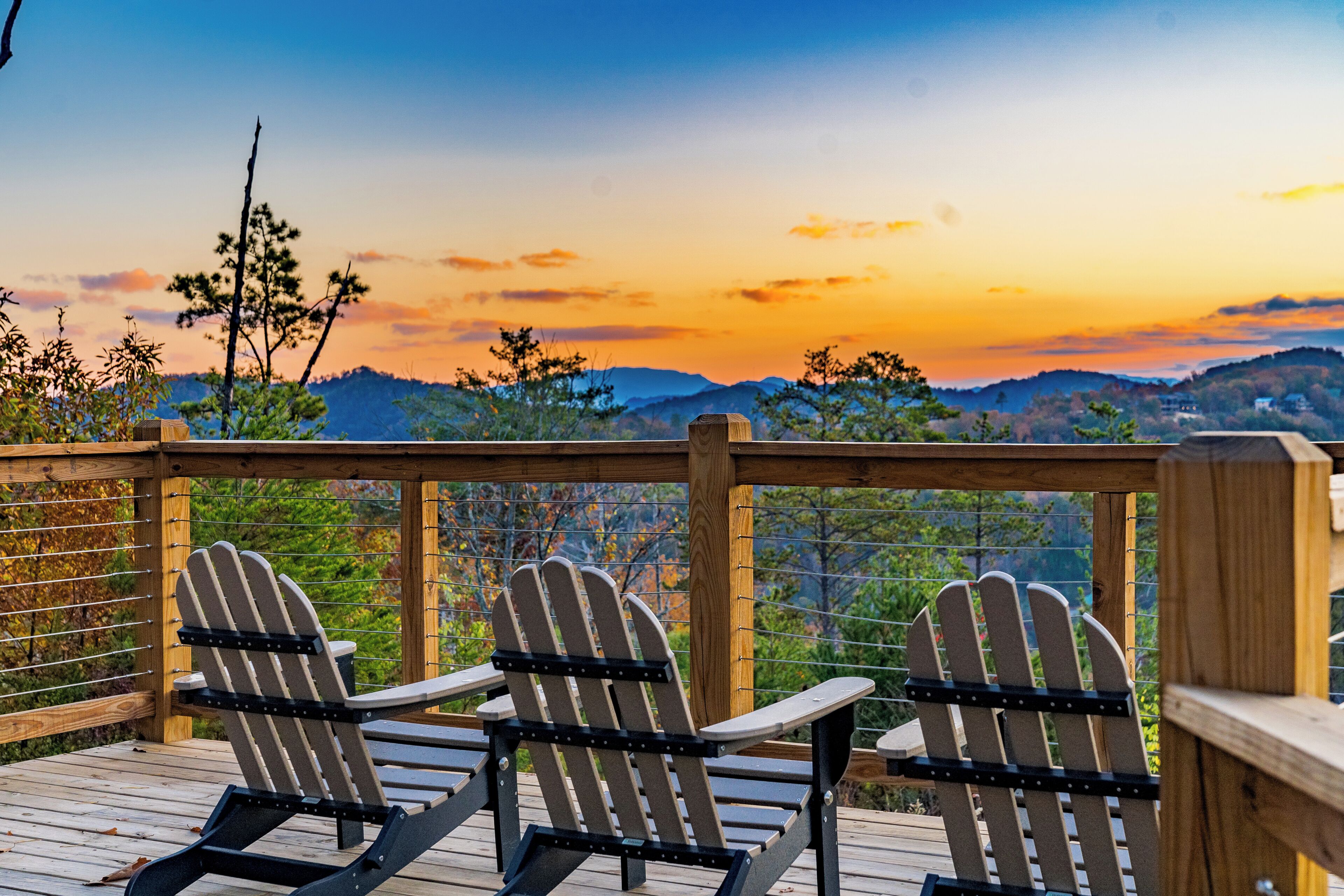 A wooden deck with Adirondack chairs overlooking a mountainous landscape during sunset.