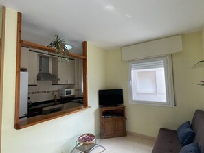 Un salon comprenant un canapé, un téléviseur posé sur un meuble en bois, une table en verre avec un saladier, et un coin cuisine avec des placards et un four à micro-ondes.