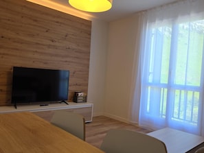 A room with a wooden wall, a flat-screen TV on a white stand, a dining table with chairs, and a large window with sheer curtains. - Modern studio near slopes - 4 guests (Aisa-Candanchú)