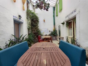 Een binnenplaats met een houten tafel en blauwe stoelen, omgeven door witte muren en groen.