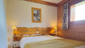 Een slaapkamer met een houten hoofdbord, twee bedlampen, een bed met een witte dekbedovertrek, een houten vloer, een raam met gordijnen en een schilderij aan de muur.