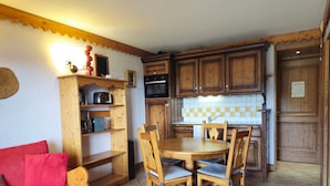 Een kleine keuken met houten kastjes, een eettafel met stoelen, een rode bank en een klein houten wandrek.