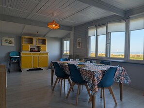 Ein Essbereich mit einem Tisch für vier Personen, einem gelben Schrank und Blick auf den Strand durch große Fenster.