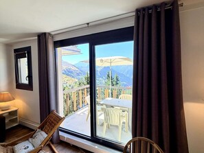 Ein Zimmer mit einer großen Glasschiebetür, die auf eine Terrasse mit Tisch und Stühlen führt, ein Sessel im Zimmer und eine Lampe auf einem Nachttisch.