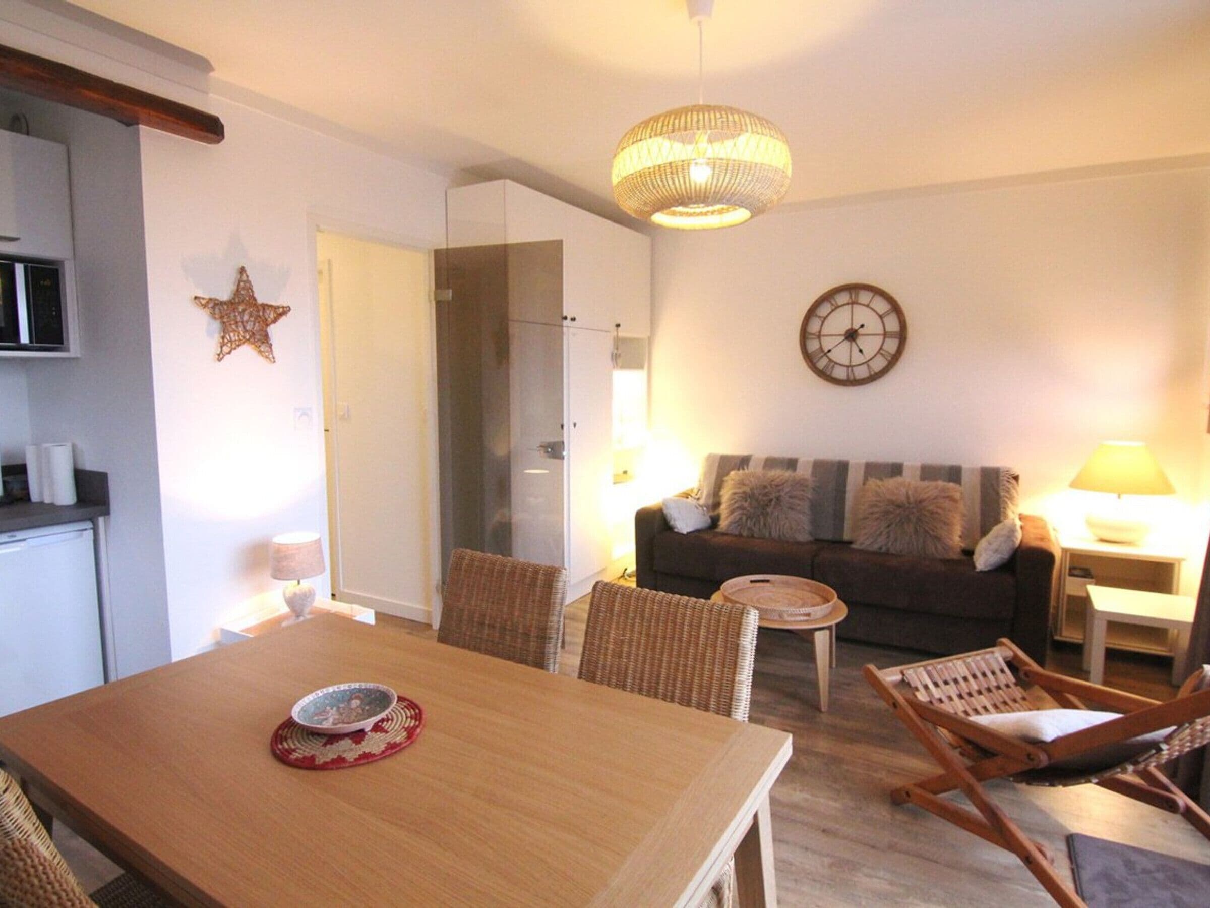 Un coin repas avec une table en bois, des chaises en osier, une petite table ronde avec une lampe, un canapé avec des coussins, une horloge murale et une décoration en forme d’étoile.