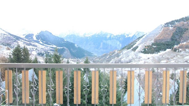 Een berglandschap met besneeuwde toppen en een balkon met een metalen hekwerk en houten latten.