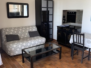 Ein Wohnzimmer mit einer Couch, einem Glastisch, einem Esstisch mit Stühlen, einer Küchenzeile mit Schränken und einem Spiegel an der Wand.