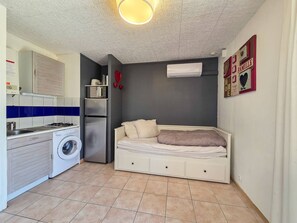 Ein kleines Zimmer mit Waschmaschine, einem Bett, einer kleinen Küchenzeile und einer wandmontierten Klimaanlage.
