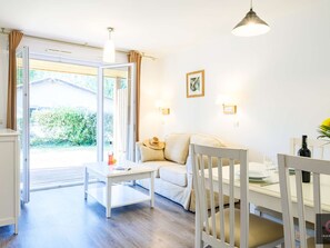 Et lyst rum med en sofa, et bord og en stol. Der er et vindue med gardiner, et billede på væggen og en lampe.