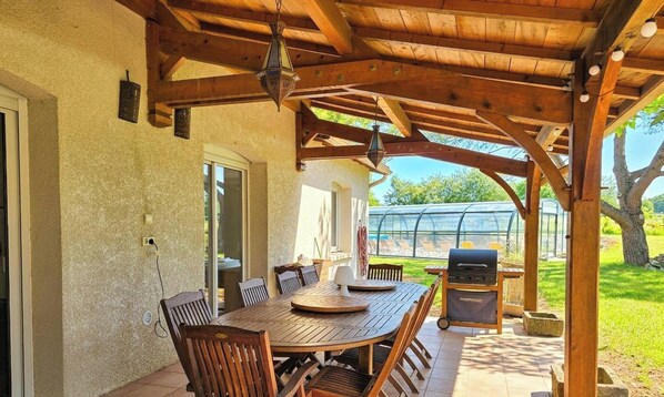 Een terras buiten met een houten tafel en stoelen, een barbecue en een overdekt gedeelte met een houten plafond.