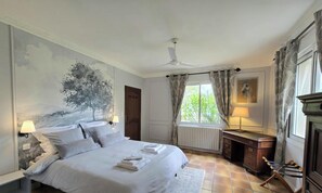 A bedroom with a bed, a desk, a radiator, a window with curtains, a ceiling fan, and a wall painting.
