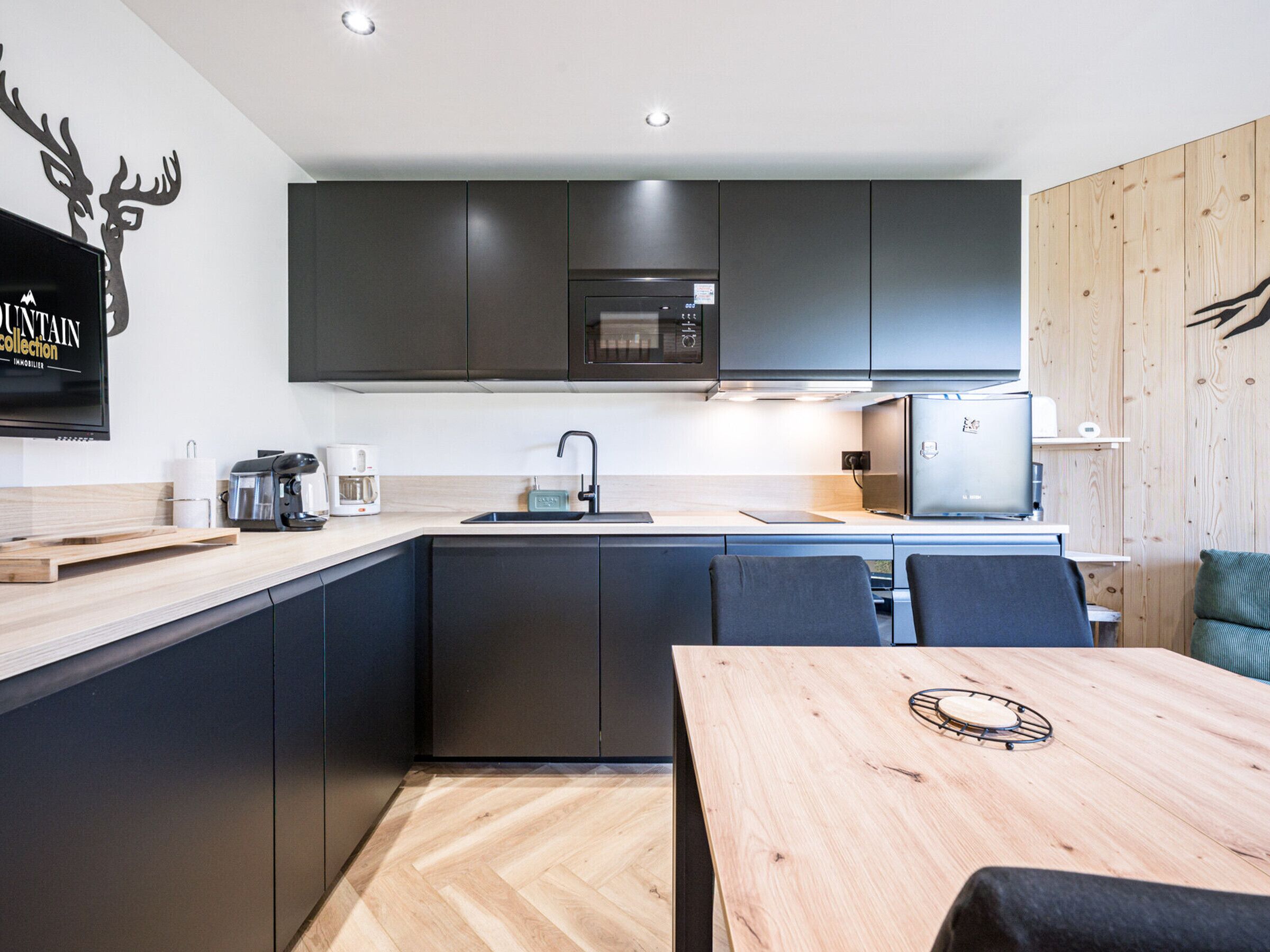 A modern kitchen with dark cabinets, a wooden dining table, and a mounted flat-screen TV.