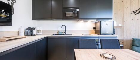 A modern kitchen with dark cabinets, a wooden dining table, and a mounted flat-screen TV.