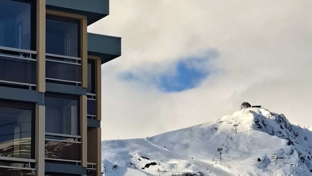 Ein modernes Mehrstöckhaus mit großen Fenstern, im Hintergrund ein verschneiter Berg und ein klarer Himmel.