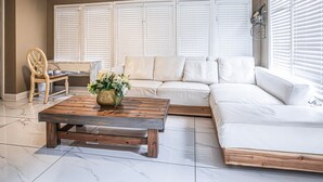 Uma sala de estar com sofá seccional, mesa de centro de madeira com um vaso, uma cadeira de madeira e uma janela com persianas.