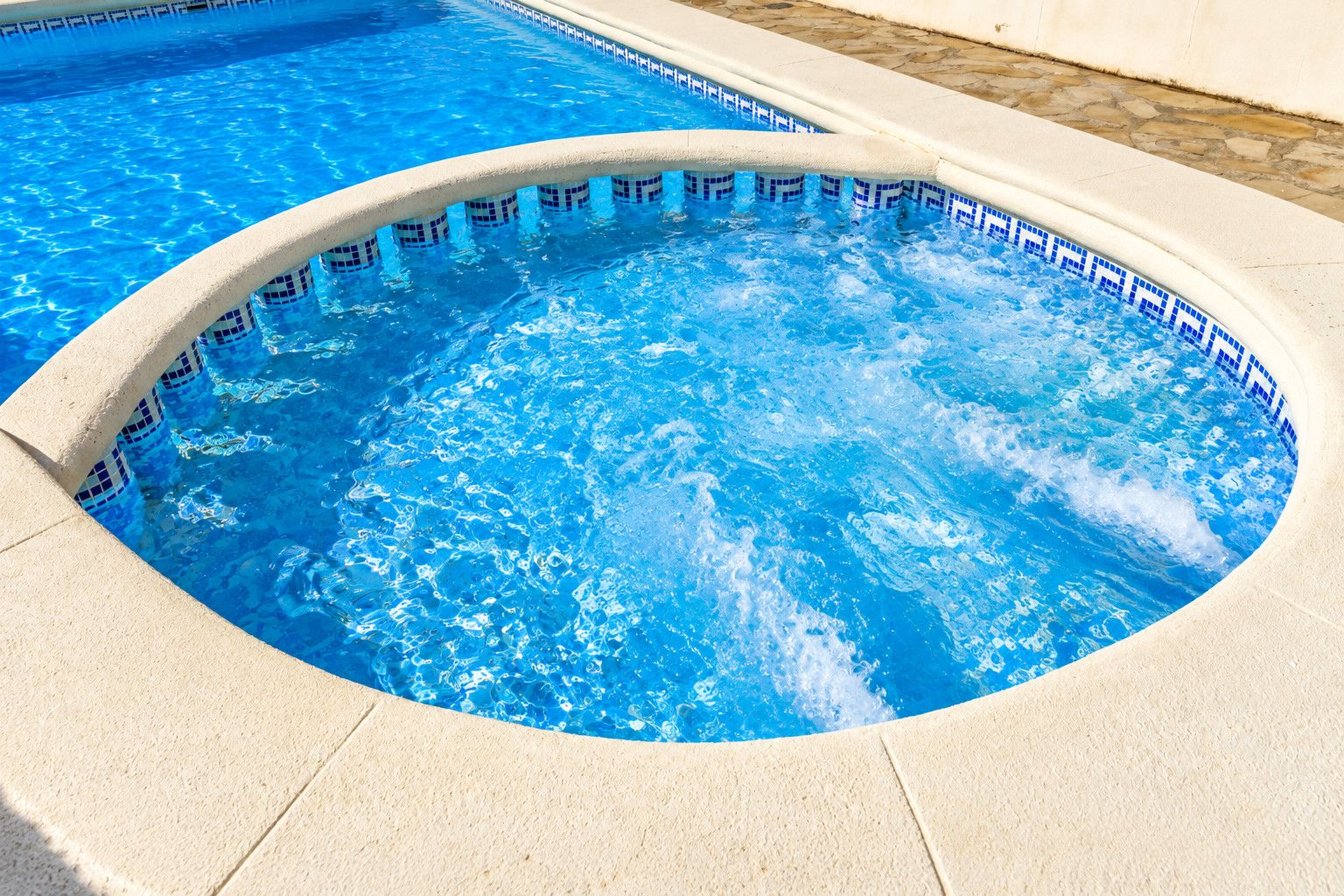 Een jacuzzi met betegelde rand, omgeven door een betonnen zwembad.