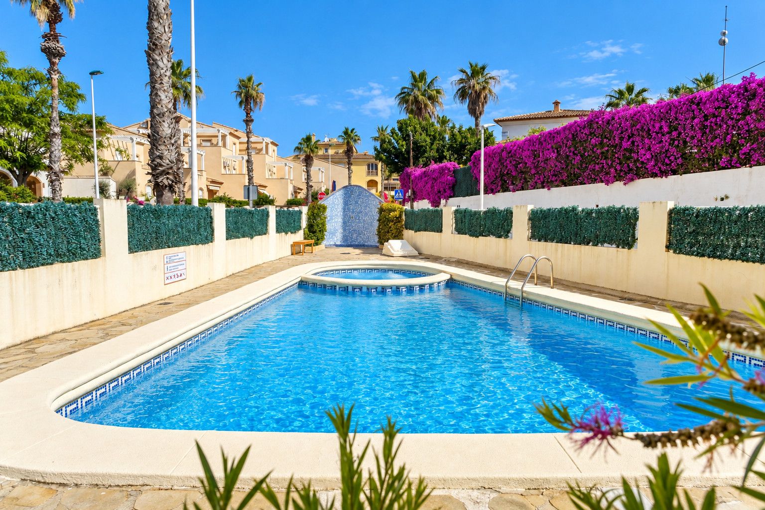 A swimming pool with a surrounding wall, palm trees, and a building in the background.