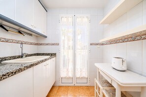 Une cuisine de taille réduite avec des meubles blancs, un plan de travail en granit, un grille-pain et une porte ornée d’un rideau en dentelle.
