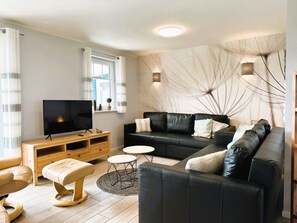 Ein modernes Wohnzimmer mit einem schwarzen Ledersofa, einer Holz-Unterhaltungseinheit mit Fernseher, einem kleinen runden Tisch und einem Fenster mit Vorhängen.