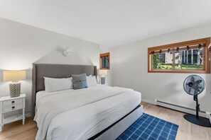 Een slaapkamer met een bed, nachtkastjes, een lamp, een ventilator en een raam met uitzicht op groen.