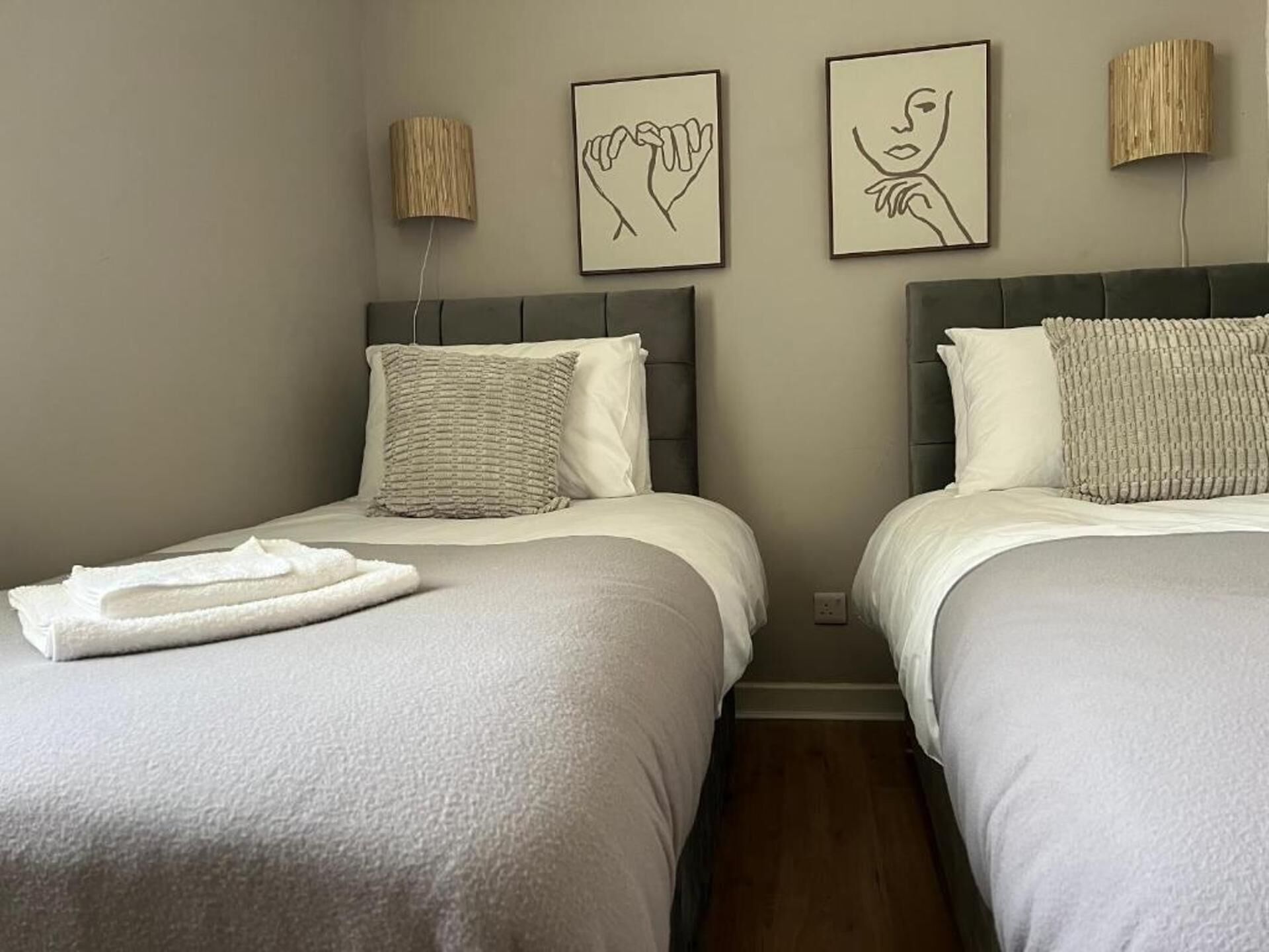 Une chambre d’hôtel avec deux lits, chacun doté d’une tête de lit grise et de linge de lit blanc. On y trouve deux lampes murales avec des abat-jour en bois, ainsi que deux œuvres d’art encadrées au-dessus des lits.