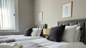 Una habitación de hotel con dos camas, una ventana con persianas, una lámpara de pared y dos cuadros enmarcados.