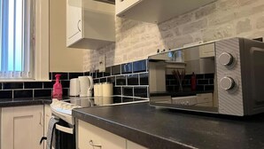 Una cocina con armarios blancos, encimera negra, microondas y ventana con persianas.