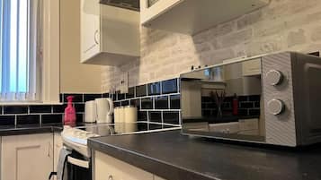 Cozinha com armários brancos, bancada preta, micro-ondas e janela com persianas.