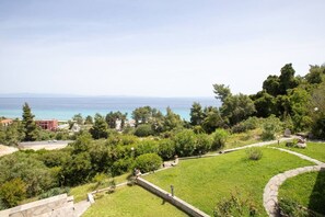 A landscaped garden with a pathway, a view of the sea, and a building in the background. - Vaya`s Maisonette by RentalsPro - Polychrono Halkidiki (Pallini)