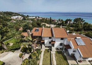 A white building with red-tiled roofs, surrounded by greenery and a view of the sea. - Vaya`s Maisonette by RentalsPro - Polychrono Halkidiki (Pallini)