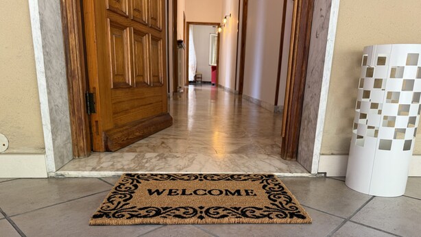 En hall med en välkomstmatta, en trädörr, en vit pelare och ett klinkergolv.