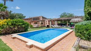 Uma piscina com deck ladrilhado, área de estar coberta e um edifício com telhado telhado ao fundo.