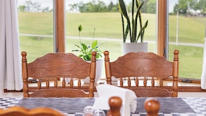 A room with two wooden chairs, a checkered mat, a white table, a potted plant, and a view of a green field through large windows.