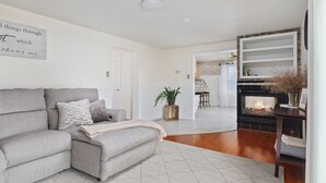 A living room with a grey sofa, a fireplace, a kitchen in the background, and a door leading to another room.
