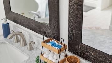 Een badkamer met een marmeren wastafel, een spiegel met een reliëfomlijsting, en een houten mandje met wattenbolletjes en blauwe wattenstokjes.