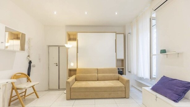 A modern, well-lit room with a beige sofa, a small wooden chair, a white table, and a window with a view. - Delightful little house to discover the truest and most popular Rome in the heart of Pigneto (Roma RM)