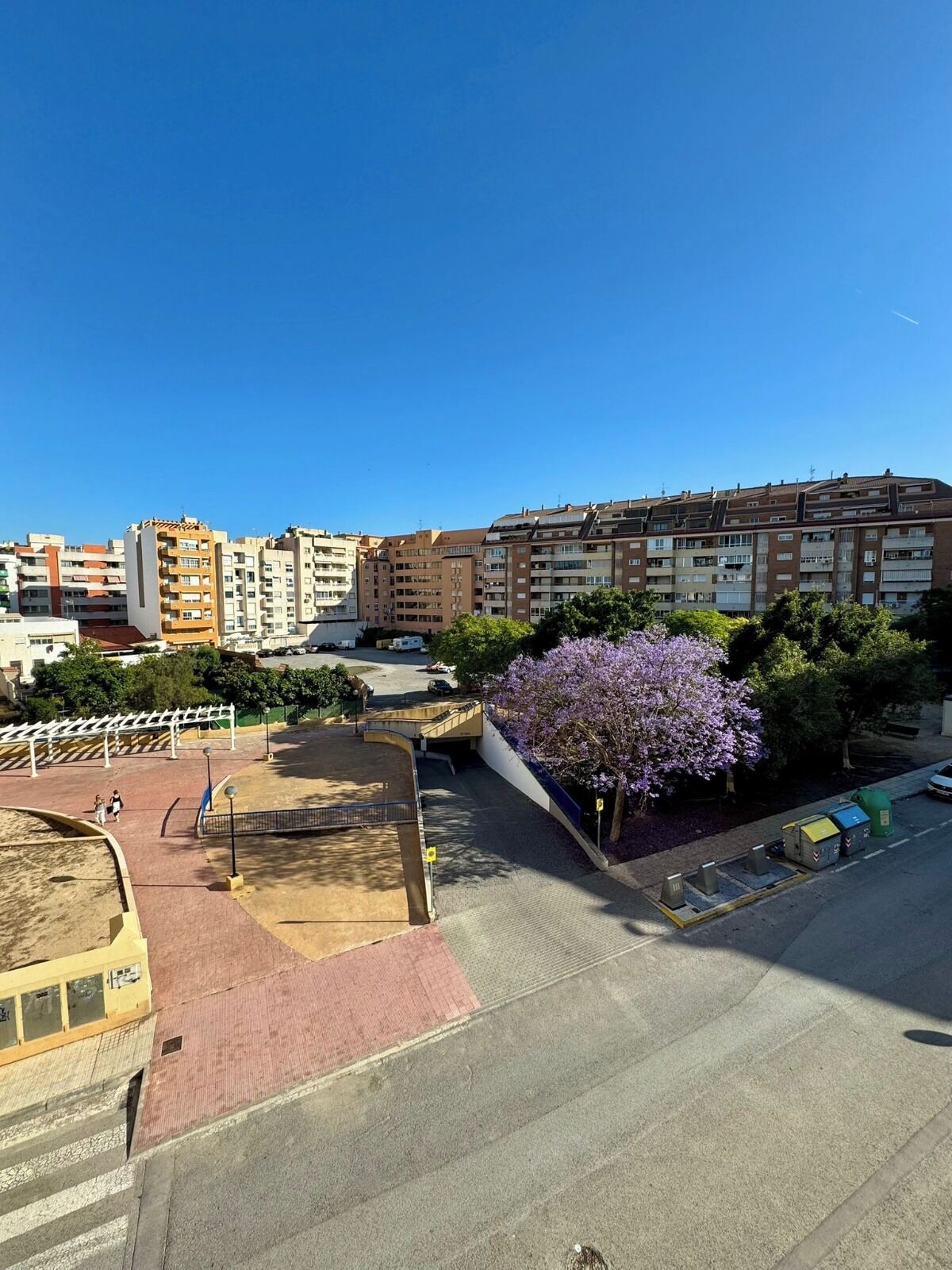 Ein Wohngebiet mit Mehrfamilienhäusern, einem Parkplatz und einem Baum mit violetten Blüten.