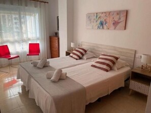 A double room with two beds, a wooden dresser, a red chair, and a painting on the wall. - Apartment very close to the beach and the subway station. (El Campello)