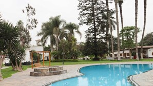 A swimming pool with a wooden deck, surrounded by palm trees and a building with a tiled roof. - w Spacious 12br Stay w Pool BBQ in Pachacamac (Pachacámac)