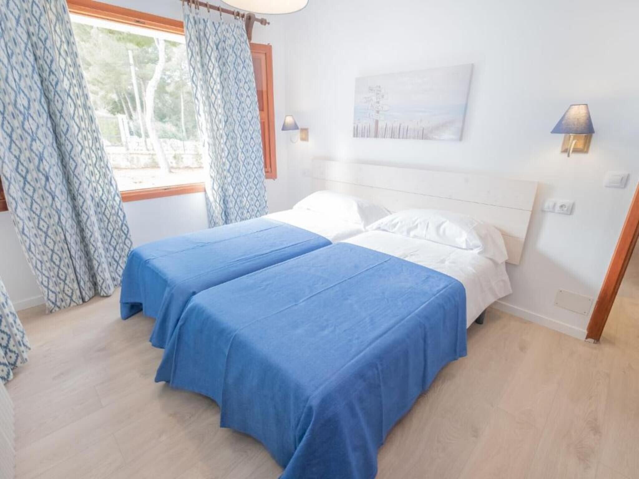 A room with two beds, a window with curtains, a wall-mounted lamp, and a painting.