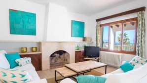 Een woonkamer met een open haard, witte banken, blauwgroene kussens, een televisie en kunstwerken aan de muren.