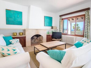 Una sala de estar con chimenea, sofás blancos, cojines color turquesa, televisión y obras de arte en las paredes.
