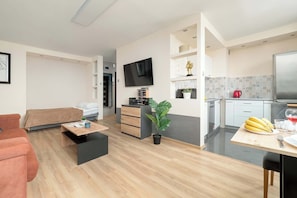 Ein modernes Wohnzimmer mit einer Couch, einem Couchtisch, einem Fernseher, einer kleinen Küchenzeile mit einem Kühlschrank und einem Esstisch mit Bananen.