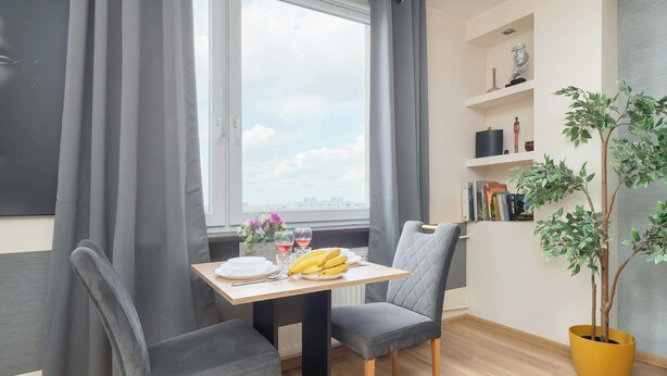 A room with a dining table, two chairs, a potted plant, a bookshelf, and a window with curtains. - Modern Studio on Piotrkowska Street City View (Łódź)
