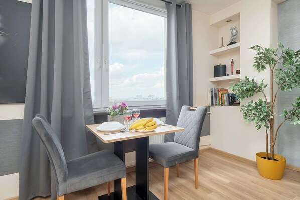Ein Zimmer mit Esstisch, zwei Stühlen, einem Blumentopf, einem Bücherregal und einem Fenster mit Vorhängen.