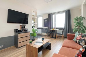 Ein modernes Wohnzimmer mit einem Flachbildfernseher, einem Holztisch, einer Couch, einem Essbereich mit Tisch und Stühlen und einem großen Fenster mit Vorhängen.