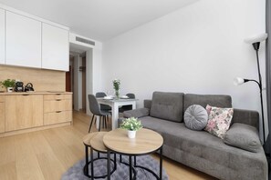 Un soggiorno moderno con un divano grigio, un tavolino rotondo con una pianta, una zona pranzo con sedie e una cucina con mobili in legno e un piano di lavoro bianco.