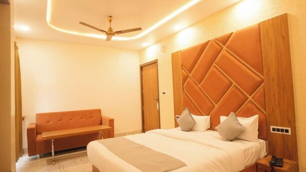 A bedroom with a bed, a sofa, a wooden headboard, a ceiling fan, and a bedside table. - Hotel Shree Karni Palace (Beawar)