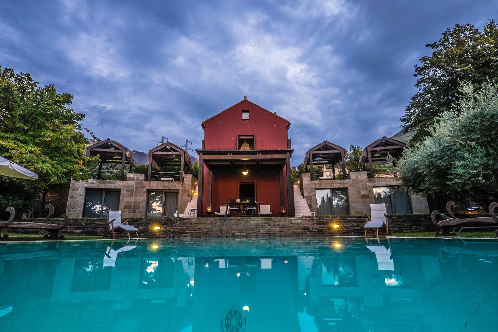 A red building with a pool in front, surrounded by trees and outdoor seating.
